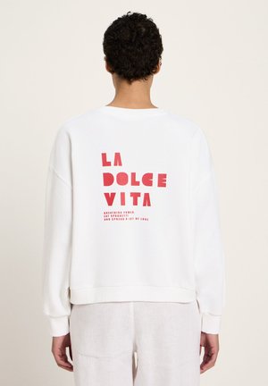 Person with curly hair wearing a white sweatshirt with red text "LA DOLCE VITA" and smaller text below on the back.