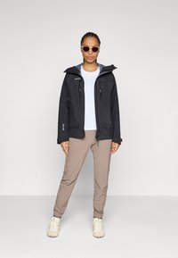 Water-resistant black jacket with two chest zippers, beige pants, and light-colored hiking shoes. Model wearing sunglasses. Neutral background.