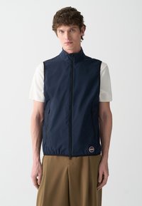 Gilet senza maniche blu navy, realizzato in materiale liscio, con zip frontale, due tasche laterali e un logo circolare vicino all'orlo.