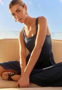 Frau mit zurückgekämmtem Haar, die ein ärmelloses navyblaues Kleid und Creolen trägt, auf einem beigen Sofa im Freien mit übergeschlagenen Beinen sitzt.