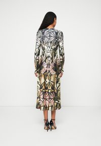 Μακριά, εφαρμοστή φόρεμα με πολύπλοκο φλοράλ σχέδιο σε πράσινο, ροζ και καφέ. Διαφανή μανίκια και ρέουσα σιλουέτα. Μαύρες ψηλοτάκουνες γόβ�ες.