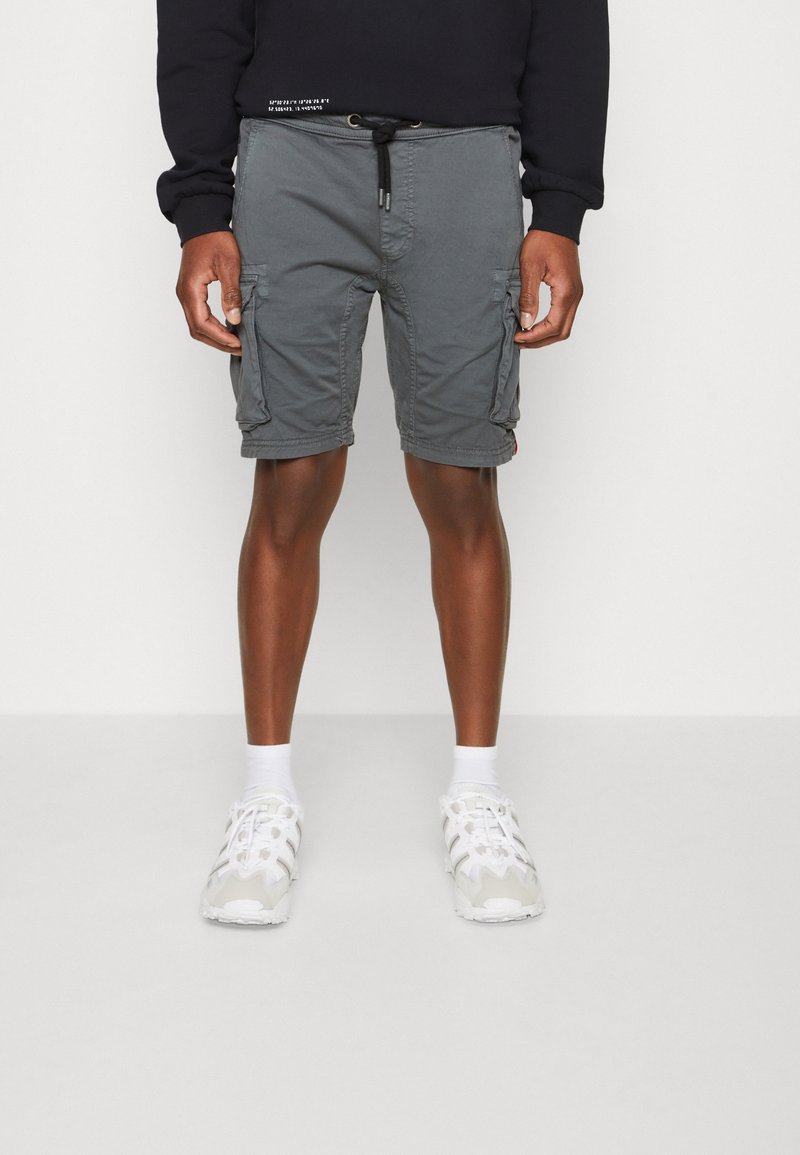 Shorts cargo gris avec poches latérales, taille à cordon de serrage et en mélange de coton. Portés avec des sneakers blanches et des chaussettes à mi-mollet.