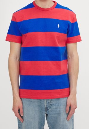 Homme portant une chemise à manches courtes larges rayures horizontales rouge et bleue avec un jean bleu clair, debout devant un fond blanc.