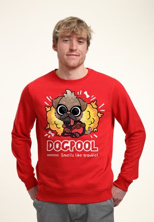 Man draagt een rode trui met een cartoon mopshond in een superheldenkostuum en tekst "DOGPOOL Ruikt naar problemen!" op de voorkant.