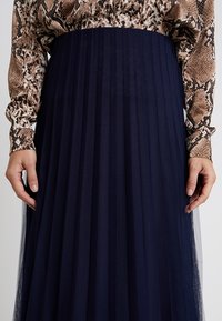 Gonna a pieghe blu navy realizzata in tessuto leggero, con un design strutturato e una vita aderente. In evidenza, mani che tengono la gonna.