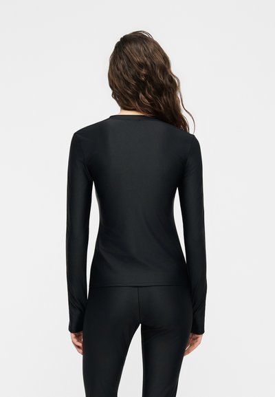 Γυναίκα με εφαρμοστή μαύρη μπλούζα μακρυμάνικη και παντελόνι, εμφανίζεται από πίσω μπροστά σε ένα μονόχρωμο ανοιχτό γκρι φόντο.