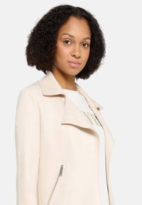 Lichtbeige jas met een reverskraag, zijritssluiting en een gladde textuur. Model draagt een wit shirt eronder.