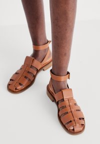 Braune Leder Sandalen mit mehreren Riemendesigns und Knöchelriemen. Niedriger Absatz, offene Zehen, mit einer glatten Textur und minimaler Hardware.
