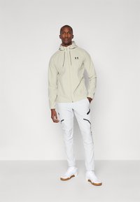 Giacca a zip color beige chiaro con cappuccio e coulisse regolabili, abbinata a pantaloni bianchi con dettagli neri e polsini. Sneakers bianche con suola marrone.
