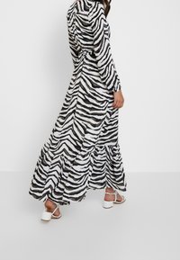 Svart och vit randig maxiklänning med flätad volang i nederkant och långa ärmar, tillverkad av lätt tyg. Har ett bälte i midjan och en avslappnad passform.