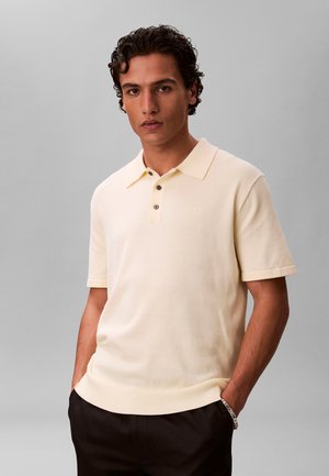 Młody mężczyzna z kręconymi włosami ubrany w kremową koszulkę polo z krótkim rękawem i czarne spodnie, stoi z rękami w kieszeniach na szarym tle.