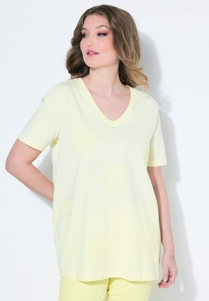 Mujer con cabello ondulado que lleva una blusa de manga corta de cuello en V de color amarillo claro y pantalones a juego, mirando de lado contra un fondo blanco.