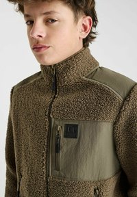 Bruine fleece jacket met olijfgroene accenten, hoge kraag, met rits sluitende zakken, gestructureerde stof en logo patch op de borst.