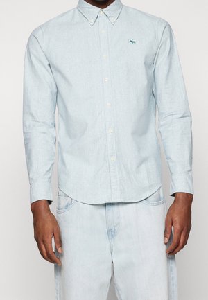Mand iført lyseblå skjorte med lange ærmer med en lille grøn elg-logo på brystet og lyseblå denimjeans.