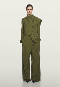 Costume sur mesure vert olive avec une veste asymétrique mettant en avant un détail boutonné et un col haut, associé à un pantalon large et ample.