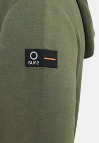 Felpa con cappuccio in tessuto verde con finitura strutturata, caratterizzata da un'etichetta nera sulla manica con logo bianco e una linea di accento arancione.