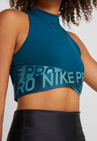 Turkusowy stanik sportowy z wysokim dekoltem, wyciętymi bokami oraz szerokim elastycznym paskiem z nadrukiem "NIKE PRO" w ciemniejszym odcieniu turkusu. Gładki, elastyczny materiał.