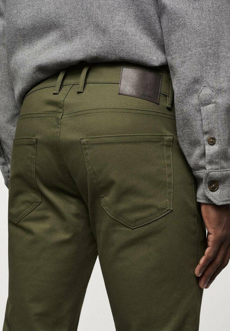 Olivgrüne Hose aus strapazierfähigem Stoff, mit zwei Gesäßtaschen und einem braunen Lederpatch am Bund. Kombiniert mit einem grauen Langarmshirt.