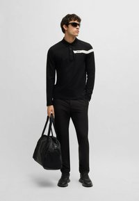 Schwarzes Langarm-Poloshirt mit einem weißen Streifen und Logo, kombiniert mit schwarzen Hosen und einer schwarzen Reisetasche. Modell trägt Sonnenbrillen.