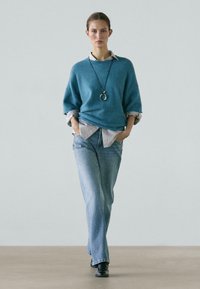 Woman walking forward wearing a teal sweater over a striped shirt, light blue jeans, black boots, and a long pendant necklace.