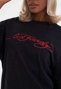 Sort bomulds t-shirt med stort rødt broderet tekst "Ed Hardy" på forsiden. Korte ærmer og en afslappet pasform.