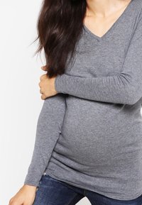 Graues Langarm-V-Shirt mit V-Ausschnitt aus weichem Material, taillierte Passform, die einen runden Bauch betont, kombiniert mit dunklen Jeans.
