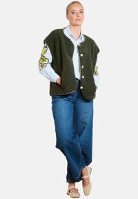 Groene fleece vest met grote knopen en zijzakken, gedragen over een blauw gestreept shirt. Gecombineerd met losse spijkerbroek en beige schoenen.