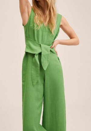 Kvinde iført en ærmeløs grøn jumpsuit med et bredt bånd bundet i taljen, poserer med den ene hånd på hoften og den anden hævet over hovedet.