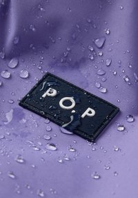 Close-up van een zwart "POP" label op paarse waterdichte stof met verspreide waterdruppels eromheen.