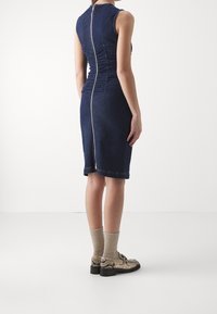 Džínové šaty s přiléhavým střihem, tmavě modré barvy, s nařasením na zadní straně a viditelným zipem. Nosené s vzorovanými botami a ponožkami.