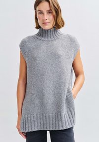 Frau in ärmellosem grauen Strickrollkragenshirt, die mit einer Hand in der Tasche vor einem schlichten Hintergrund steht.
