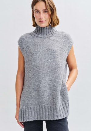 Femme portant un pull col roulé sans manches en tricot gris, se tenant avec une main dans la poche contre un fond uni.
