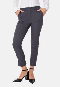 Pantaloni grigi su misura con tasche laterali, chiusura a bottone, gambe affusolate e una texture liscia, abbinati a scarpe nere con tacco.