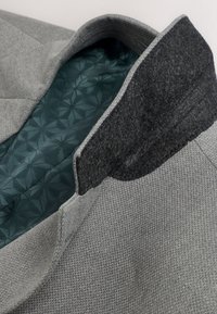 Col de manteau en tissu gris texturé avec sous-col doux gris foncé et doublure intérieure visible à motif géométrique bleu sarcelle.