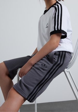 Osoba siedząca na białym krześle, ubrana w białą koszulkę Adidas z czarnymi paskami oraz szare spodenki Adidas z czarnymi paskami.