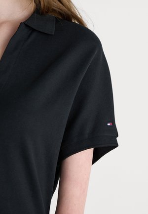 Polo noir en tissu texturé avec un col et des manches repliées. Doté d'une petite étiquette logo aux couleurs rouge, blanc et bleu.