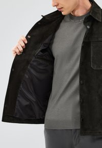 Giacca scura in suede con un esterno strutturato, chiusura a bottone e un rivestimento nero liscio. Indossata sopra un maglione grigio lavorato a maglia.