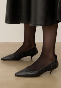 Escarpins en cuir noirs avec un bout pointu, une garniture froncée autour de l'ouverture et un petit talon, portés avec des collants noirs transparents.