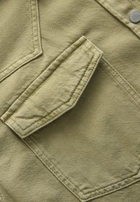 Khakifarbenes Gewebe mit strukturierter Oberfläche, versehen mit einer einzelnen aufgenähten Tasche auf der Vorderseite, die eine spitze Klappe und einen metallischen Druckverschluss hat.