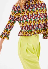 Farverig trykt bluse med rynket kant, med geometriske former i rød, gul, grøn og blå, parret med klare gule bukser.