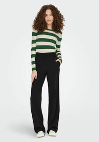 Gestreepte ribbentrui in groen en roomkleur, zwarte wijde broek en witte sneakers; figuur staat pose met één hand in de broekzak.