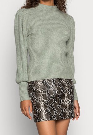 Femme portant un pull côtelé vert clair avec des manches bouffantes et une jupe courte marron à imprimé serpent.