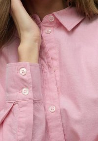 Camisa de botón de color rosa claro con cuello, botones similares a perlas y una tela suave y texturizada. Los puños tienen tres botones cada uno.