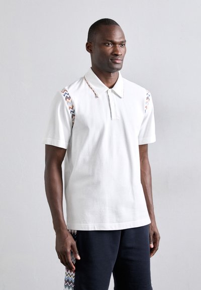 T-shirt blanc à manches courtes et col. Présente des motifs en zigzag colorés sur les épaules. Tissu en coton doux.