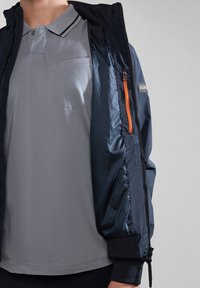 Marineblå bombeflyjakke med en tekstureret overflade og orange lynlåsdetaljer over en grå polo-shirt med brystlomme og stribet krave.