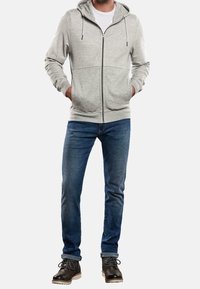 Sweat à capuche gris zippé avec capuche, poches kangourou et poignets côtelés, associé à un jean bleu et des bottes noires à lacets.