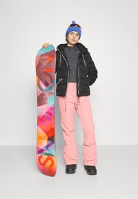Schwarze Pufferjacke, graue Midlayer, rosa Schneehose, schwarze Stiefel, blauer Beanie. Hält ein mehrfarbiges Snowboard mit "SNOW" Grafik.