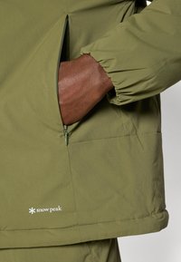 Olive green jacket with a side pocket, elastic cuffs, and a small white "snow peak" logo at the hem. Lightweight fabric with a smooth texture.