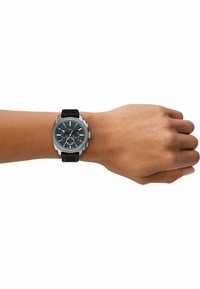 Sort kronografur med tekstureret grå urskive og sølvdetaljer på et sort silikonearmbånd, båret på en lysbrun arm.