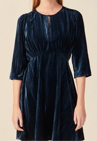 Vestido de terciopelo azul marino con escote redondo, mangas tres cuartos y un detalle de ojal en la parte delantera. Presenta un diseño plisado y una falda acampanada.